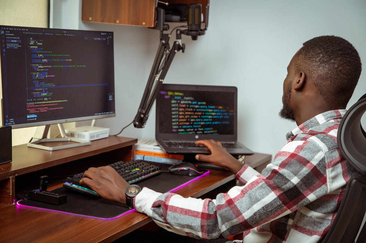 An African man coding on a desktop and laptop in a Nairobi office setting, showcasing modern technology.